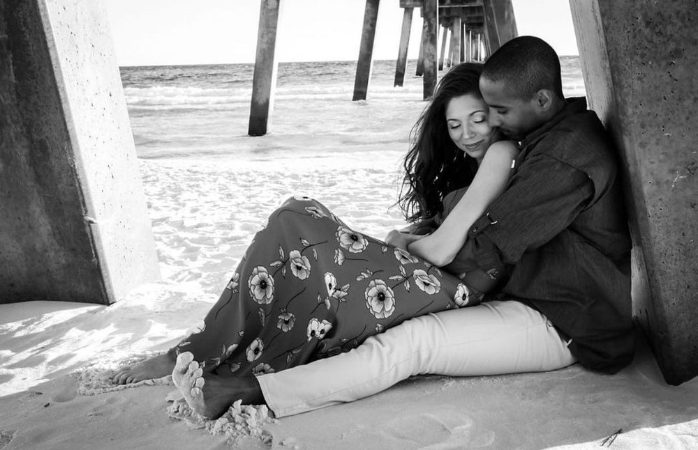 Romantic couple sitting together under beach pier in black and white engagement photo.
