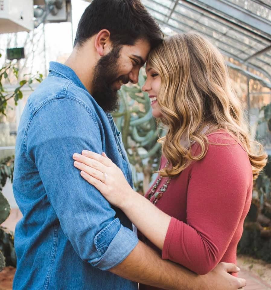A couple shares a tender moment smiling at each other while embracing inside a lush greenhouse.
