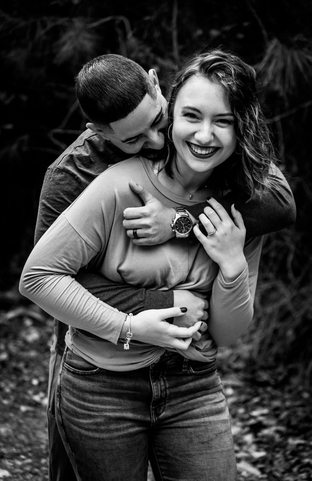 Black and white photo of a happy couple embracing outdoors, woman laughing joyfully as man hugs her from behind.