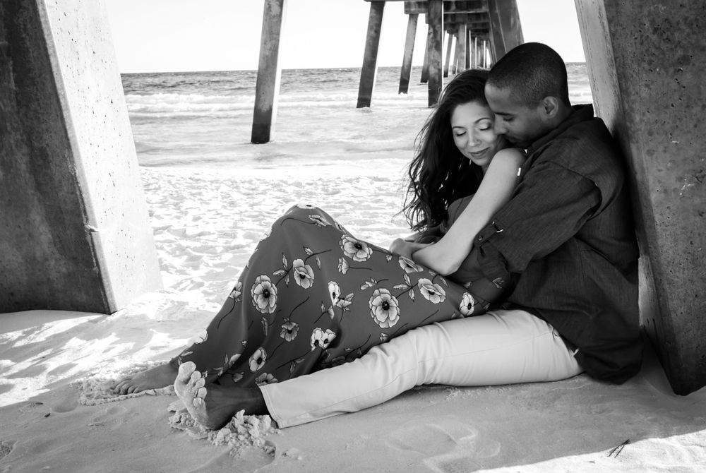 Romantic couple cuddling under a beach pier in a black and white photo on sandy shore.