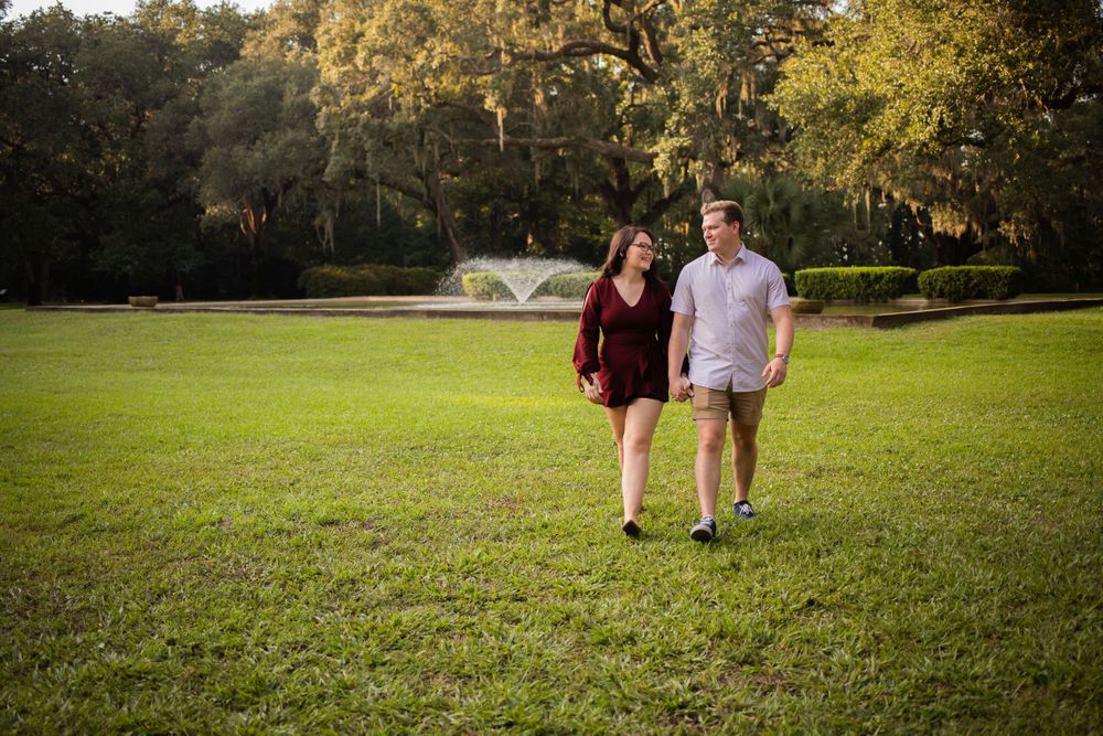 Happy couple walking hand in hand across a lush green lawn with a fountain and oak trees behind them.