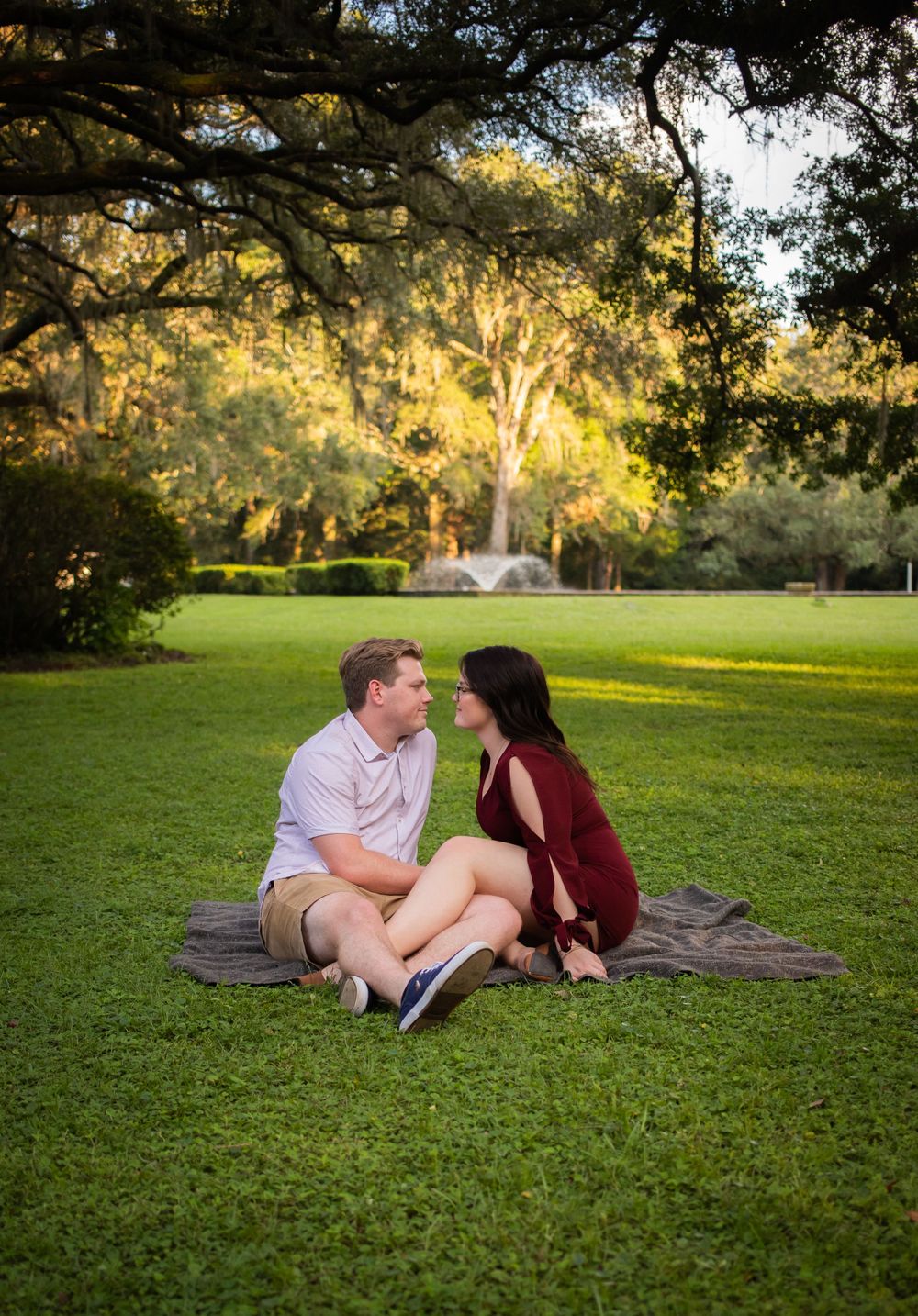 Couple sitting on a blanket in a sunlit park with large oak trees during a romantic engagement session.