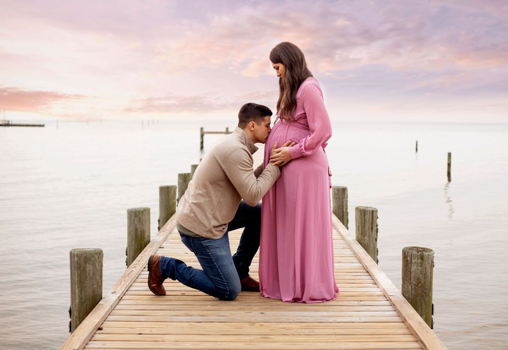 Man kneeling to kiss pregnant woman's belly on a wooden pier at sunset.