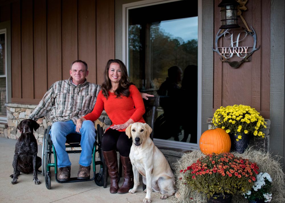 Couple with two dogs on a decorated autumn porch, man in wheelchair beside fall flowers.