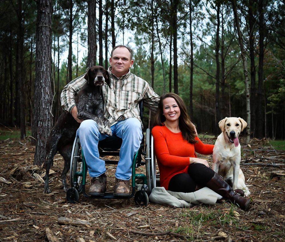 Couple with two dogs posing in a pine forest, man in wheelchair smiling outdoors.