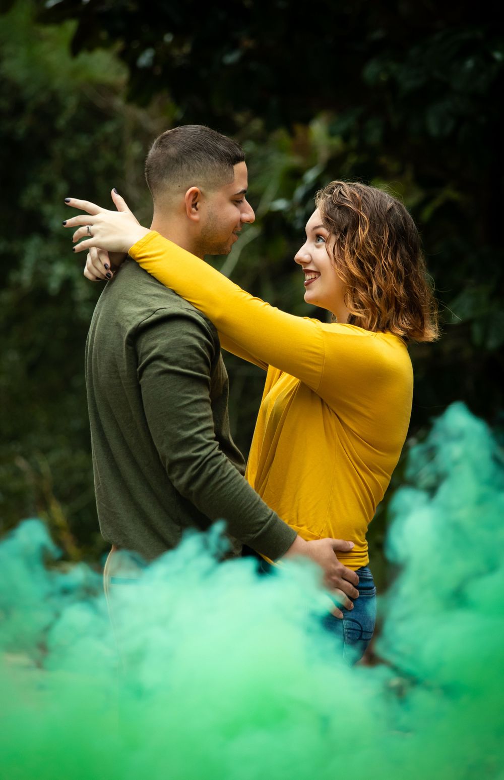 Smiling couple embraces warmly surrounded by green smoke in a lush outdoor forest setting.