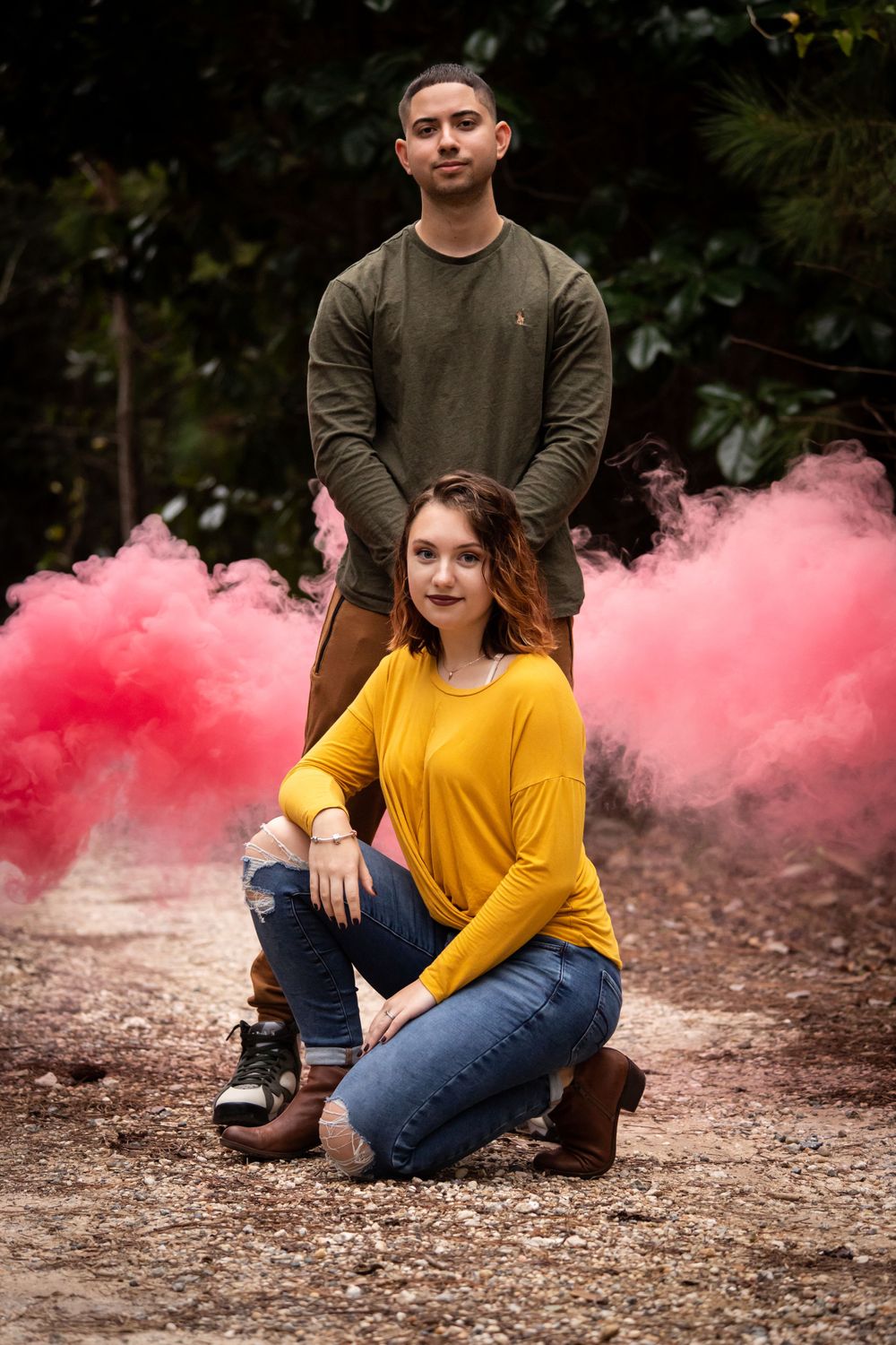 Couple poses dramatically on a gravel path surrounded by vibrant pink smoke in a forest setting.
