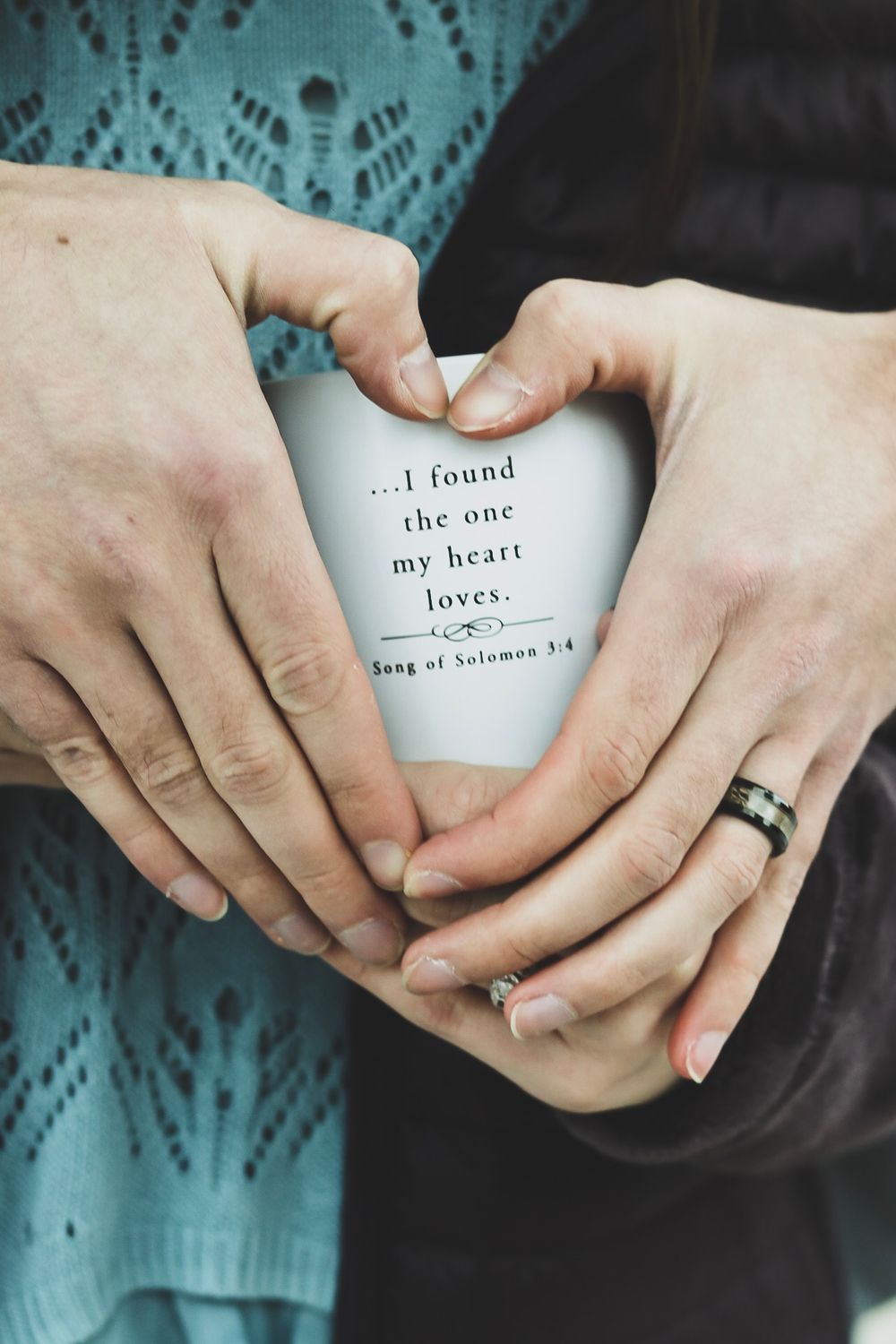 A couple forms a heart shape with their hands around a mug inscribed with Song of Solomon 3:4 verse.
