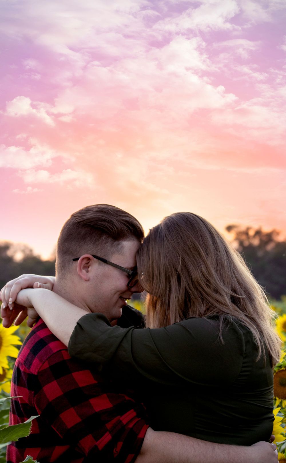 Romantic couple embracing in sunflower field at sunset with pink and purple sky background.