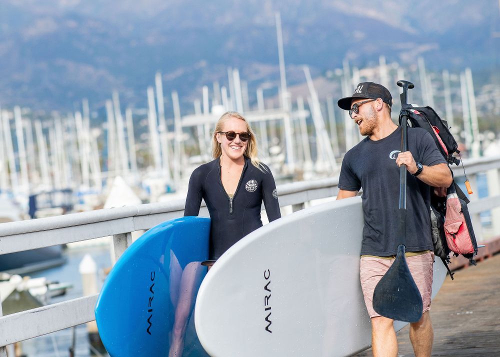 action shot of man and woman walking together at the santa barbara harbor carrying paddle boards