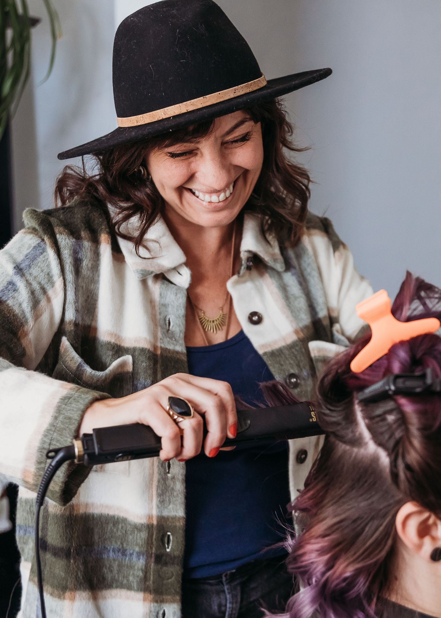 A hairstylist wearing a plaid jacket and black hat working with styling tools.