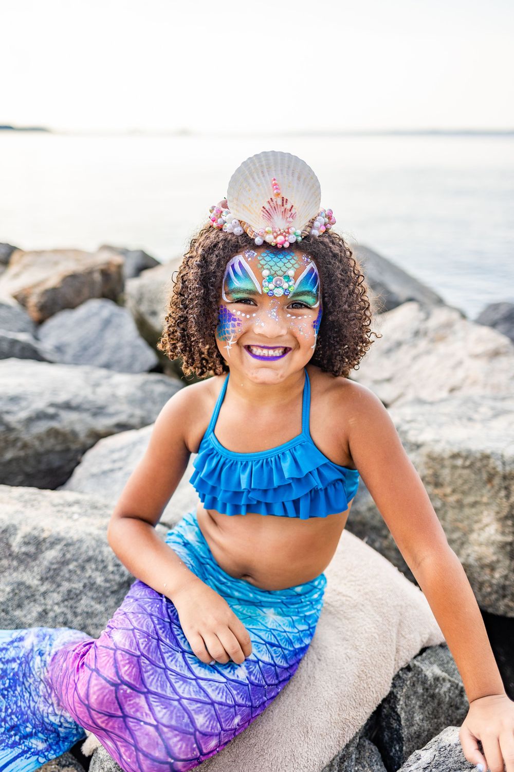 Colorful mermaid costume with face paint and shell crown against rocky beach backdrop.