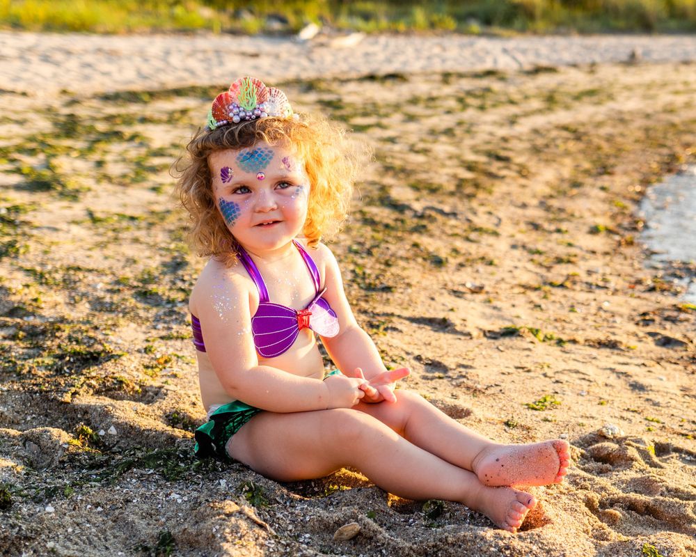 Magical mermaid photoshoot on sunny beach with sparkling face paint and crown.