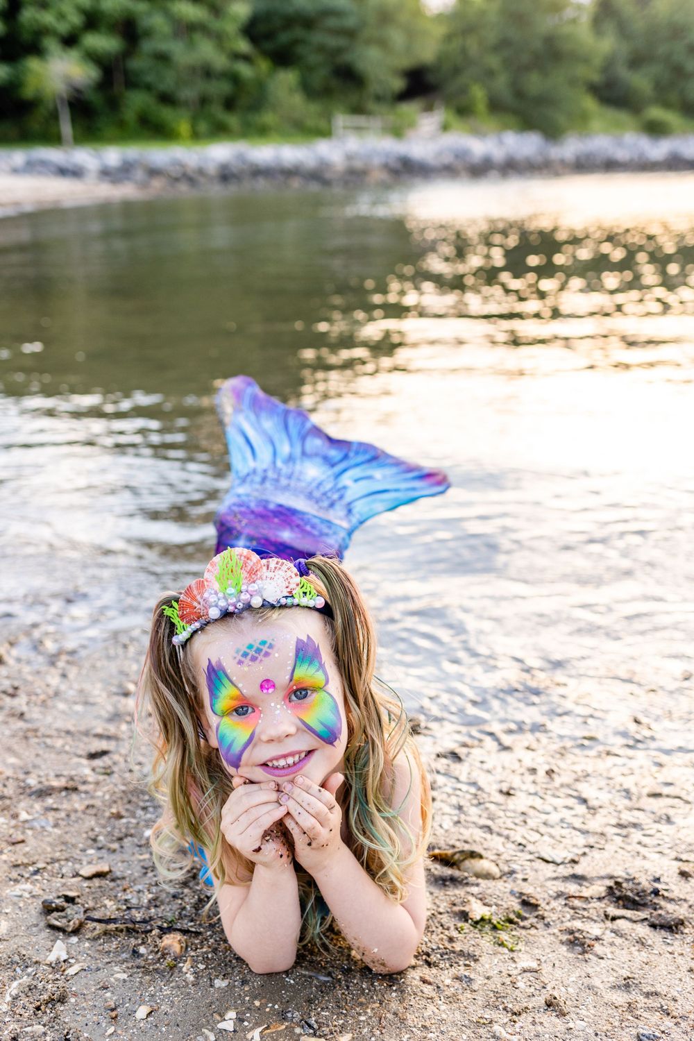 Whimsical mermaid pose with colorful face paint and shell crown by waterfront.