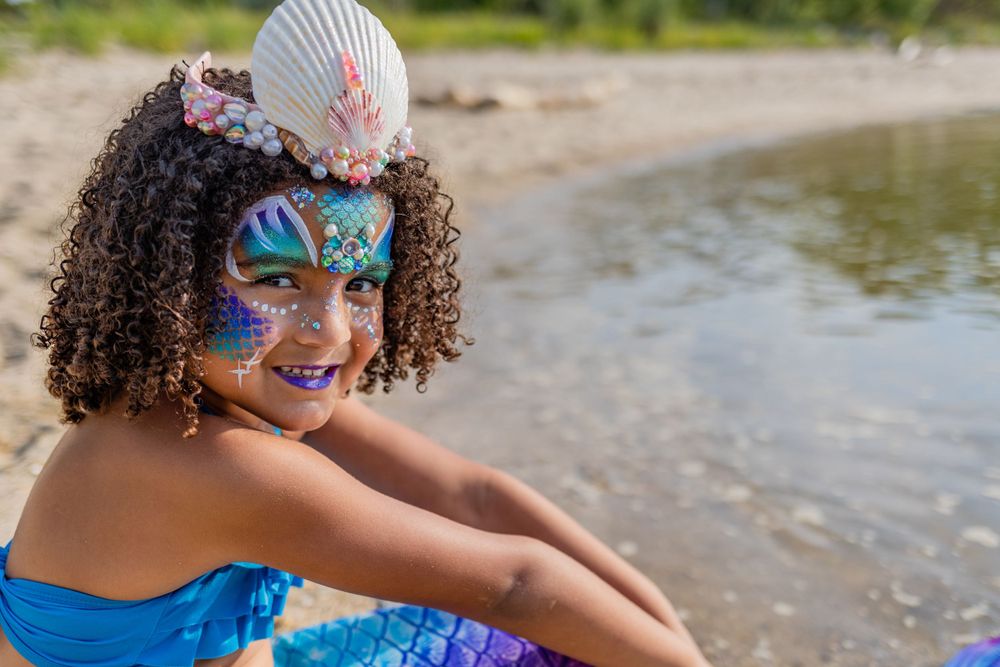 A mermaid with shell crown and face paint poses on sunny beach.