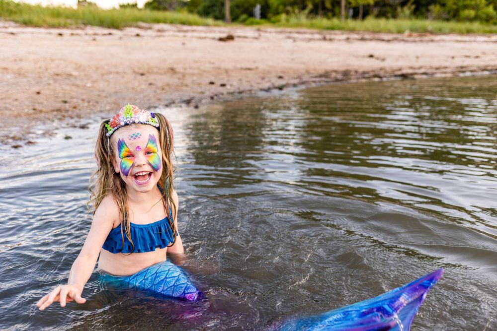 Playful mermaid splashing in shallow water with bright face paint and blue tail.