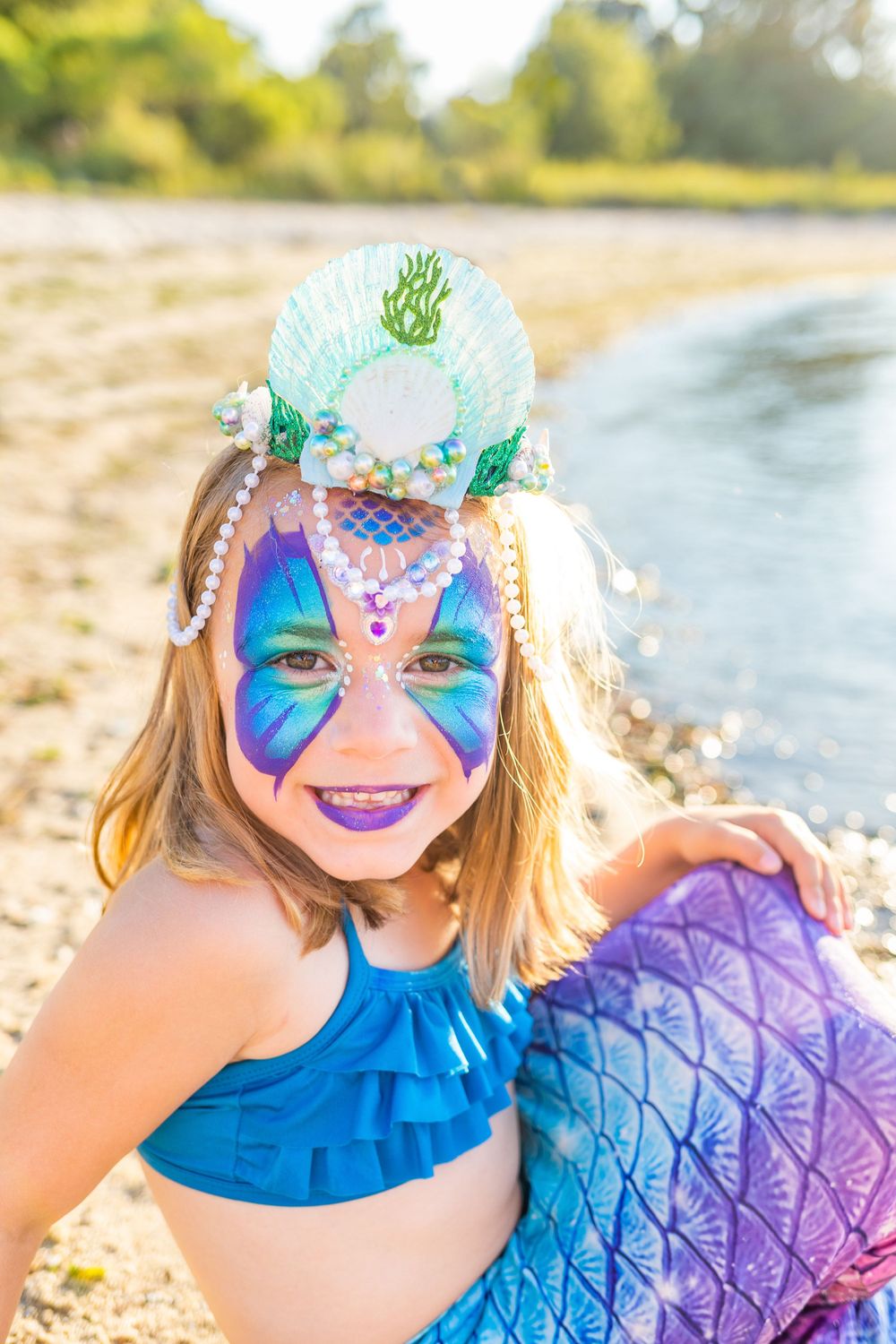 Vibrant face paint and seashell crown complement blue mermaid costume at beach.