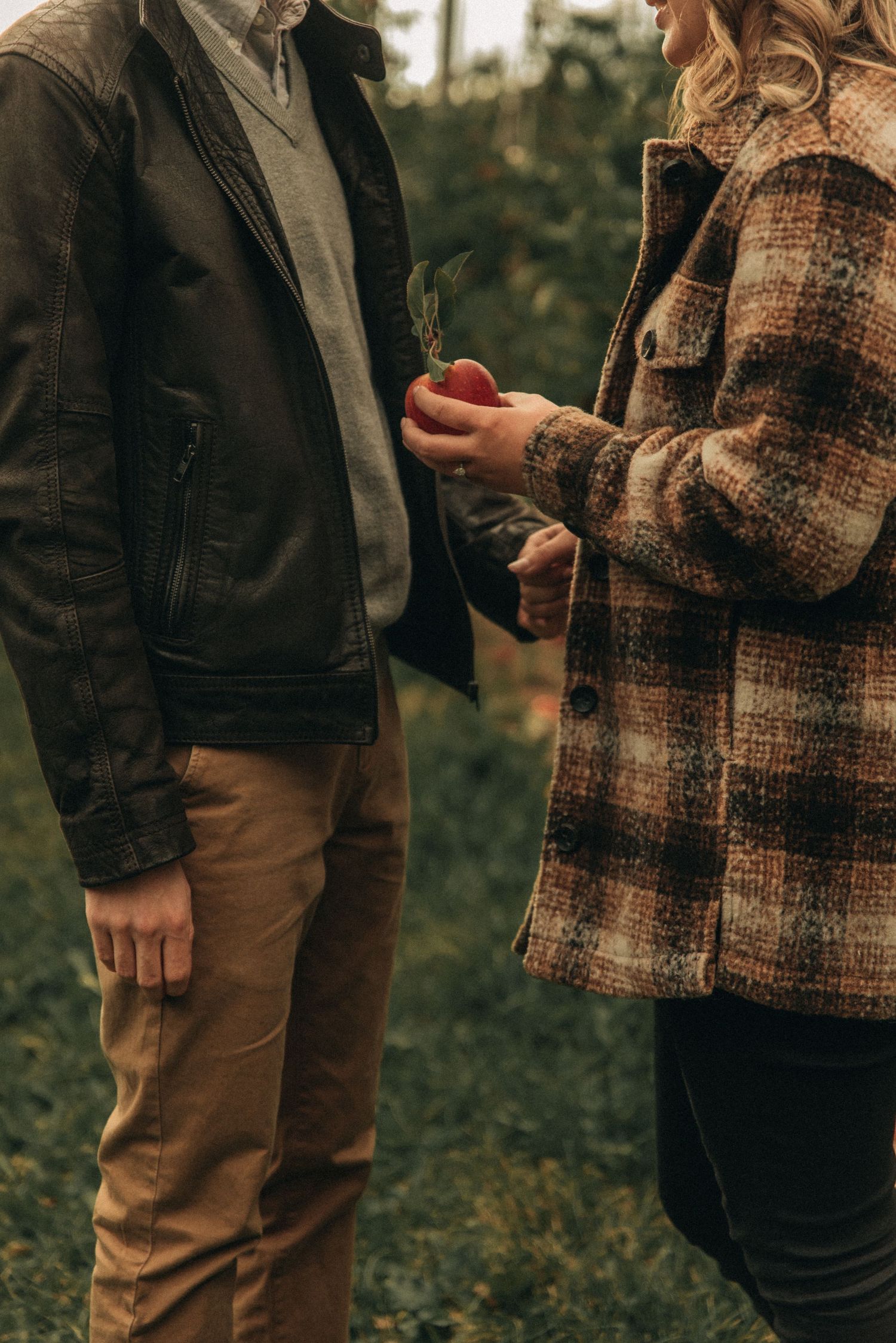 Two people in autumn clothing share a moment while holding an apple in an orchard.