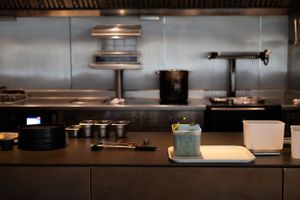 A blue cocktail sits on a dark counter in a dimly lit restaurant kitchen with stainless steel equipment visible.