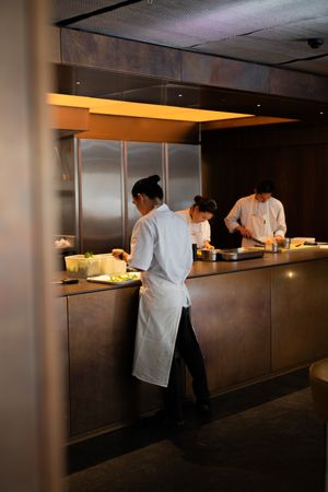 Chefs in white uniforms work at a dimly lit restaurant counter with warm overhead lighting.