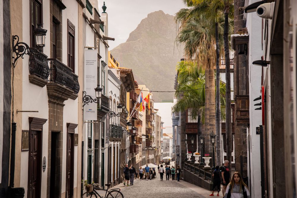 Charming cobblestone street lined with white buildings, palm trees, and a dramatic mountain backdrop.