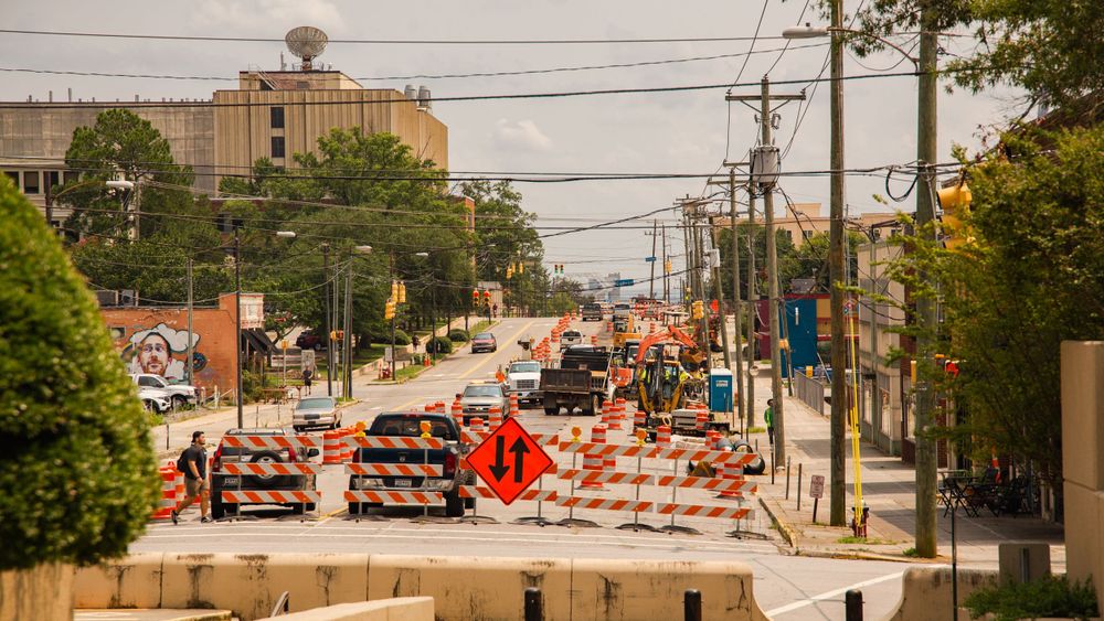 Urban road under construction with orange traffic barriers and work vehicles on a busy city street.