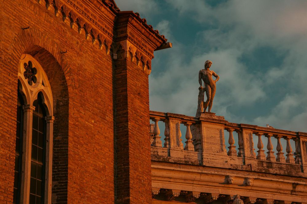 Classical statue on ornate balustrade of historic red brick building bathed in golden sunset light.