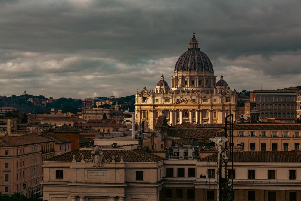 St. Peter's Basilica dome rises majestically over Vatican City rooftops under moody dramatic clouds.