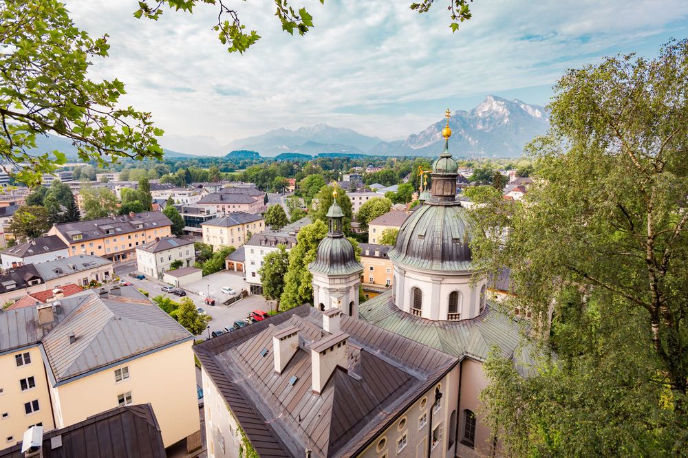 Aerial view of Salzburg old town featuring baroque church domes and mountain backdrop.