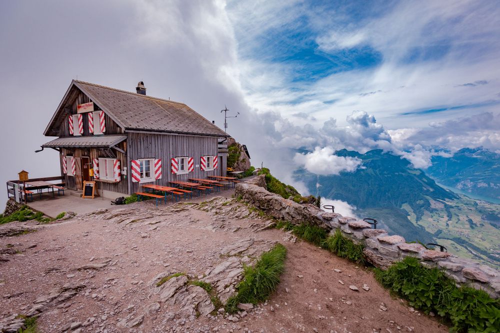 Rustic mountain hut on rocky summit with dramatic cloudy sky and valley views below.