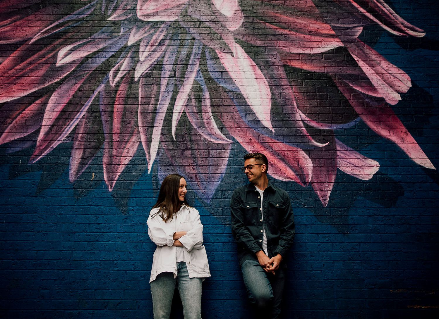 Romantic couple poses against blue wall with pink floral mural creating dreamy engagement photo backdrop. Worcestershire Wedding photographe