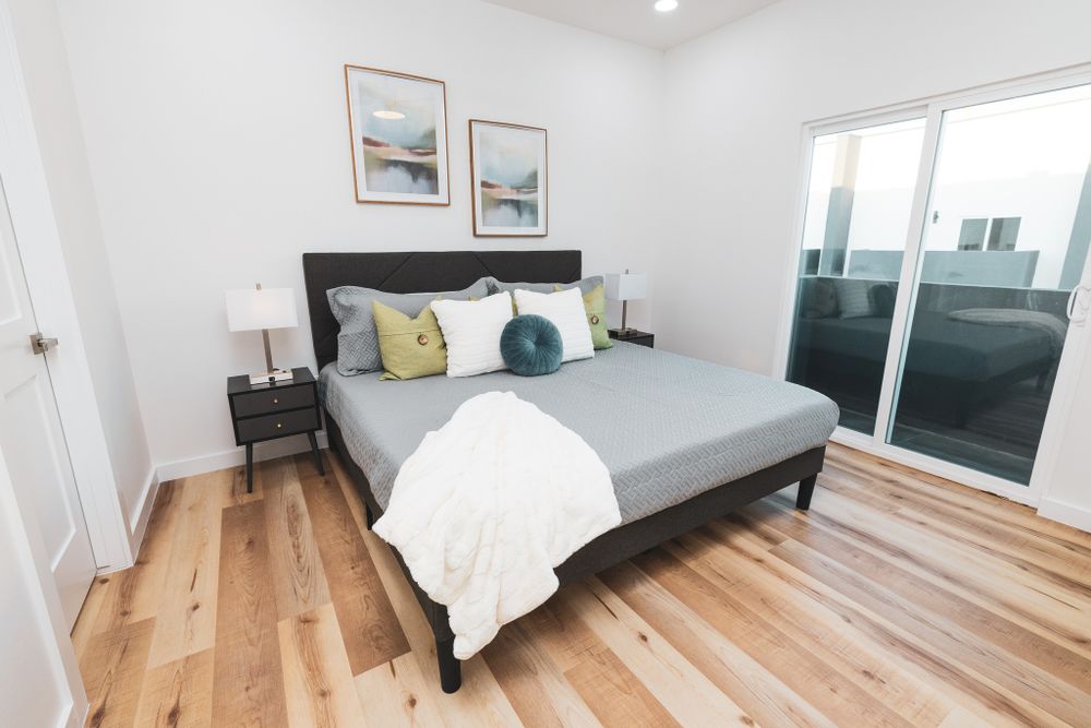 Modern bedroom with wooden floors, grey bedding, coastal artwork, and sliding glass doors.