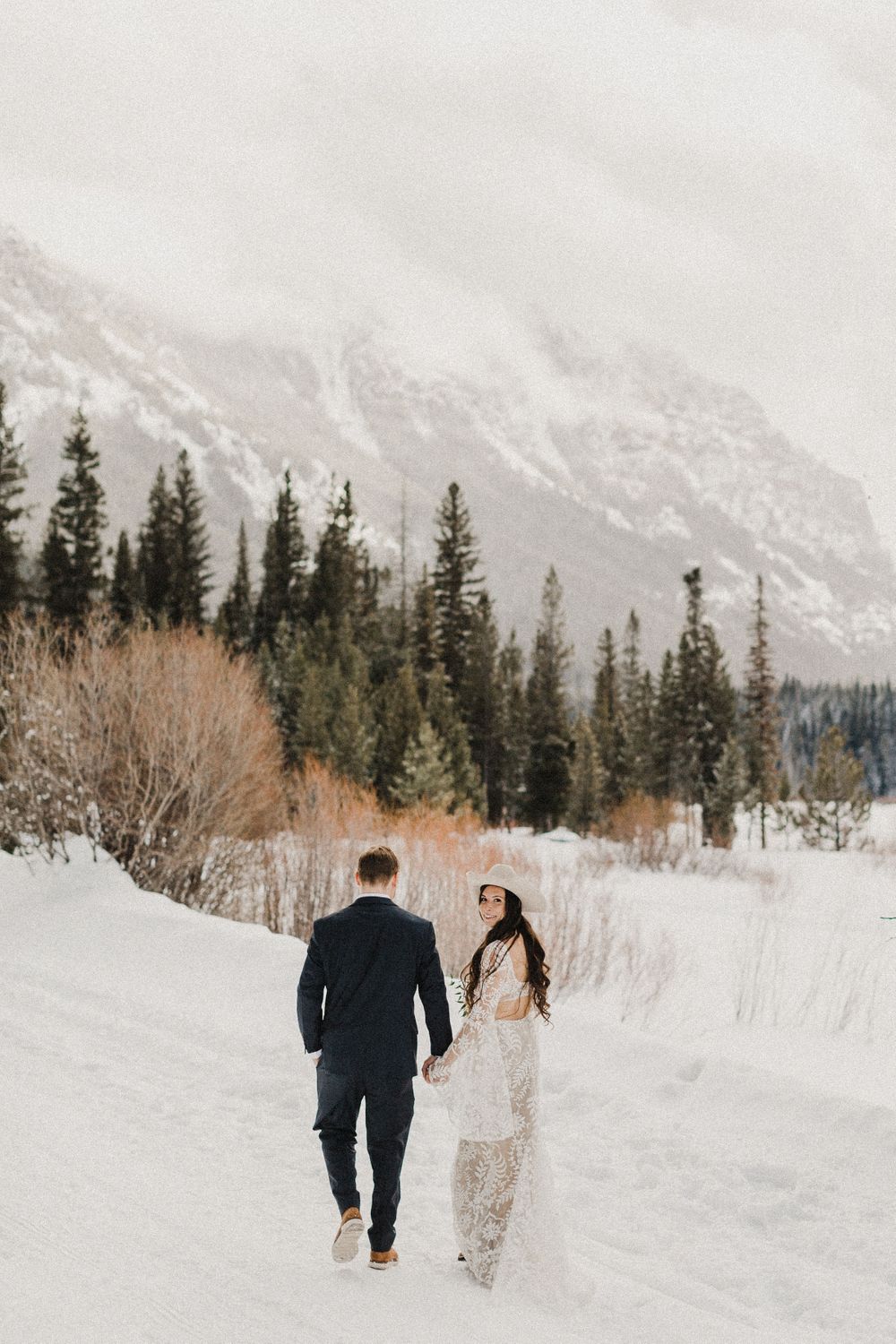 Couple walking through snowy mountain landscape with evergreen trees.