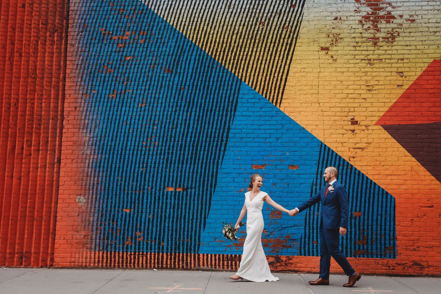 A newlywed couple walks hand in hand against a vibrant geometric wall mural of blue, red, and yellow colors in DUMBO Brooklyn.