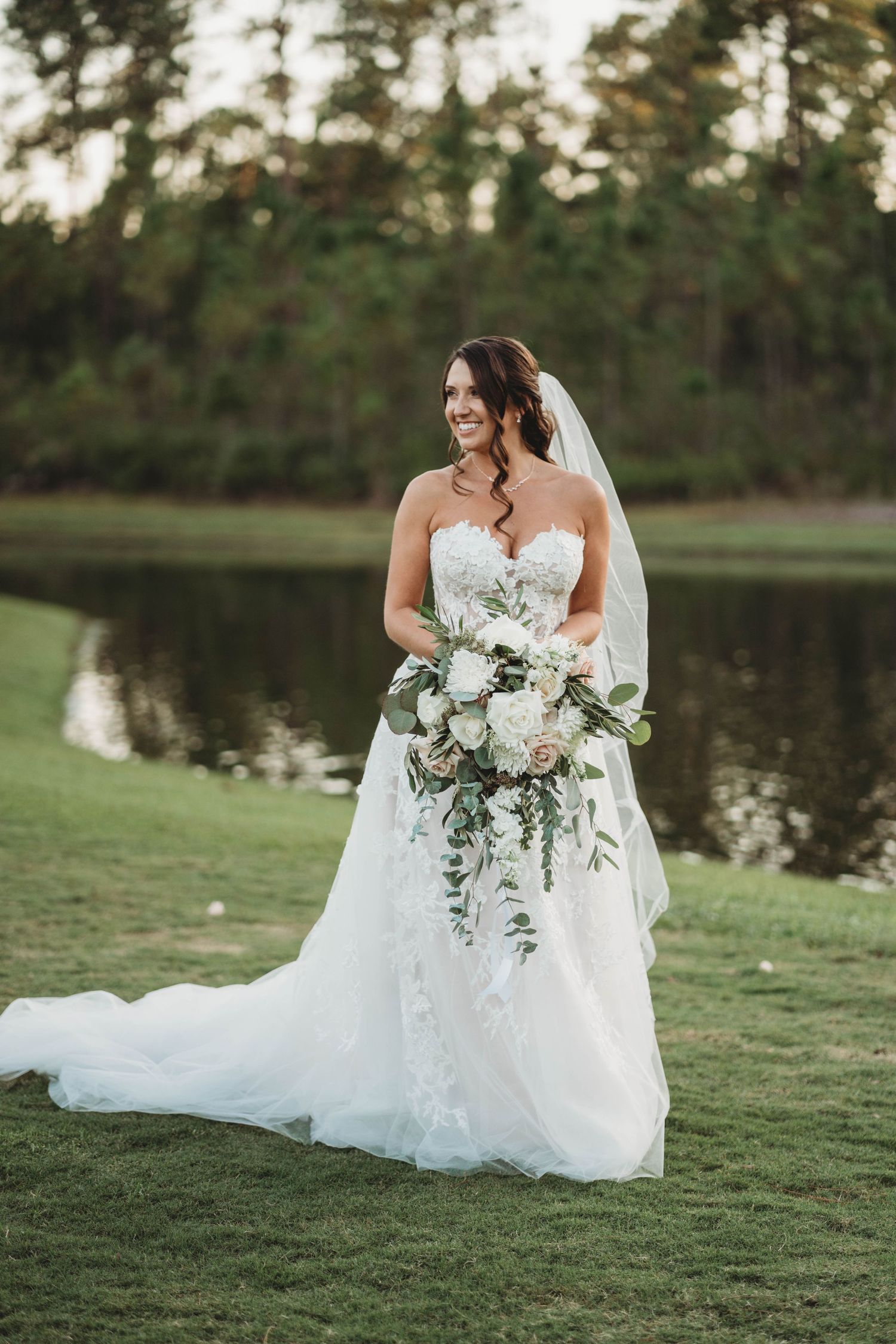 Bride in strapless white gown poses with cascading white bouquet by lakeside on lush green lawn.