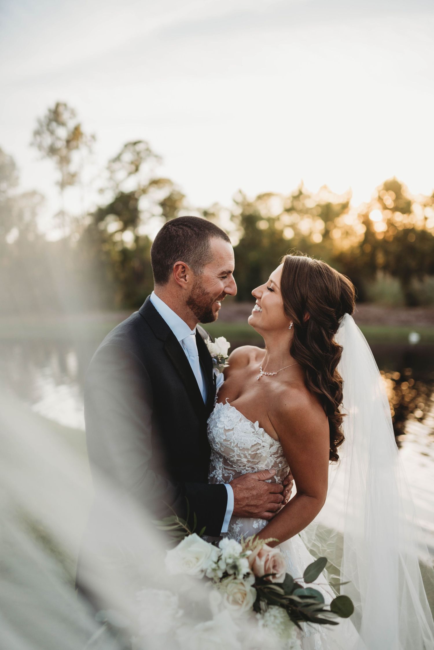 Romantic sunset portrait of bride and groom embracing by lakeside.
