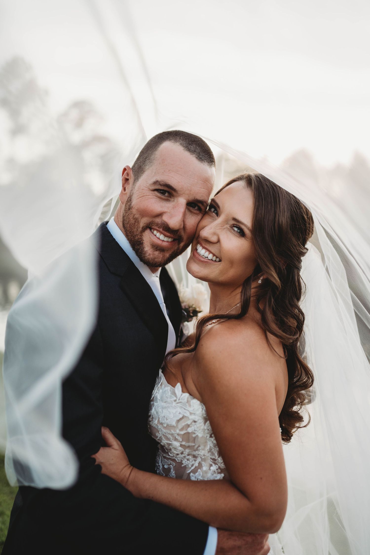 Happy newlyweds smiling together under bridal veil during outdoor portrait session.