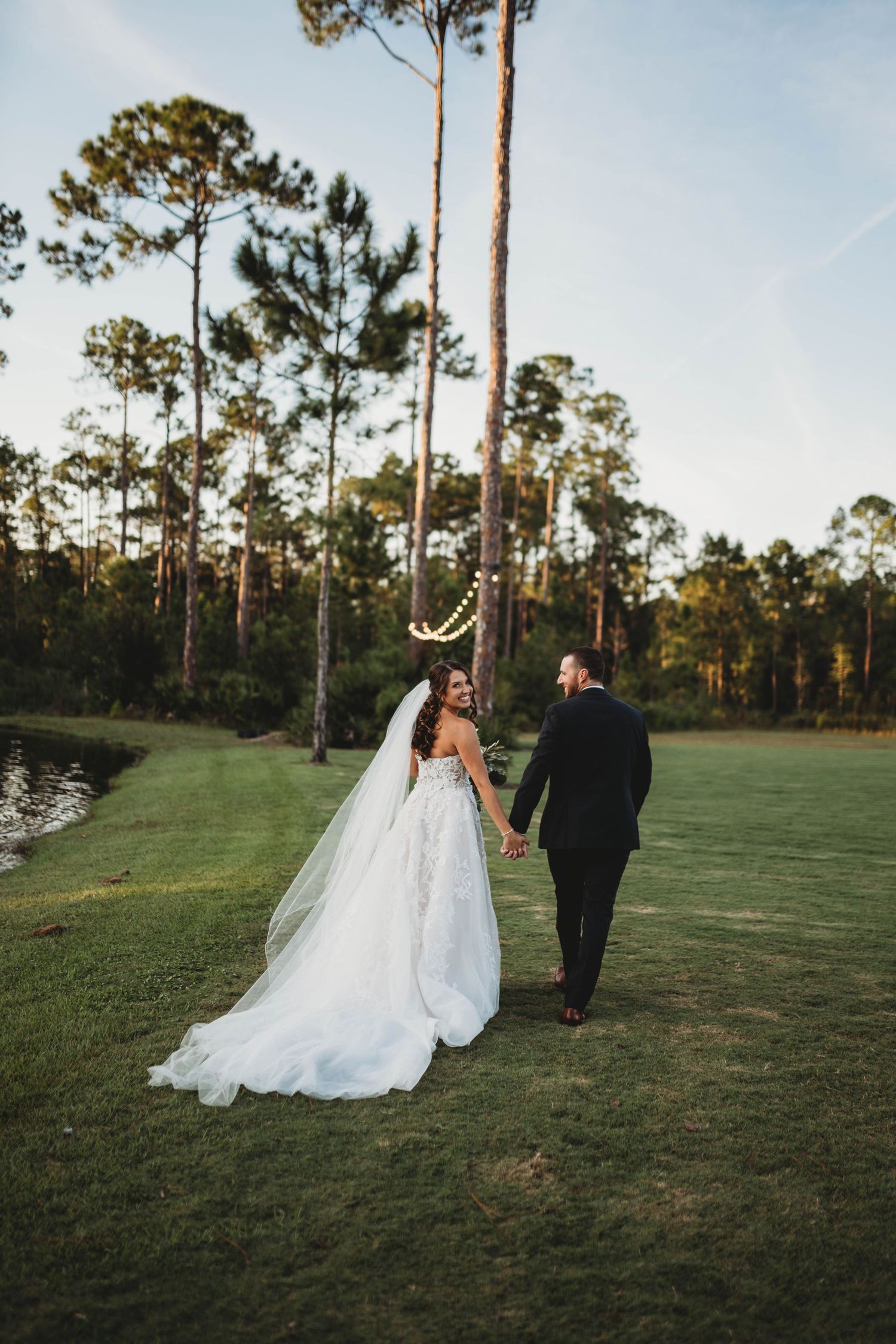 Bride and groom walking hand in hand on golf course with tall pine trees in background.