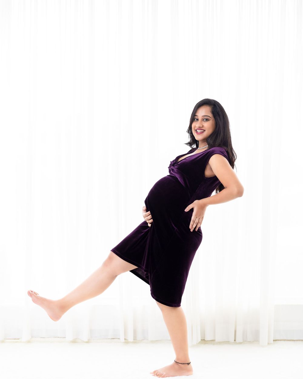 A maternity photo of someone in a purple velvet dress striking a playful pose against a white backdrop.