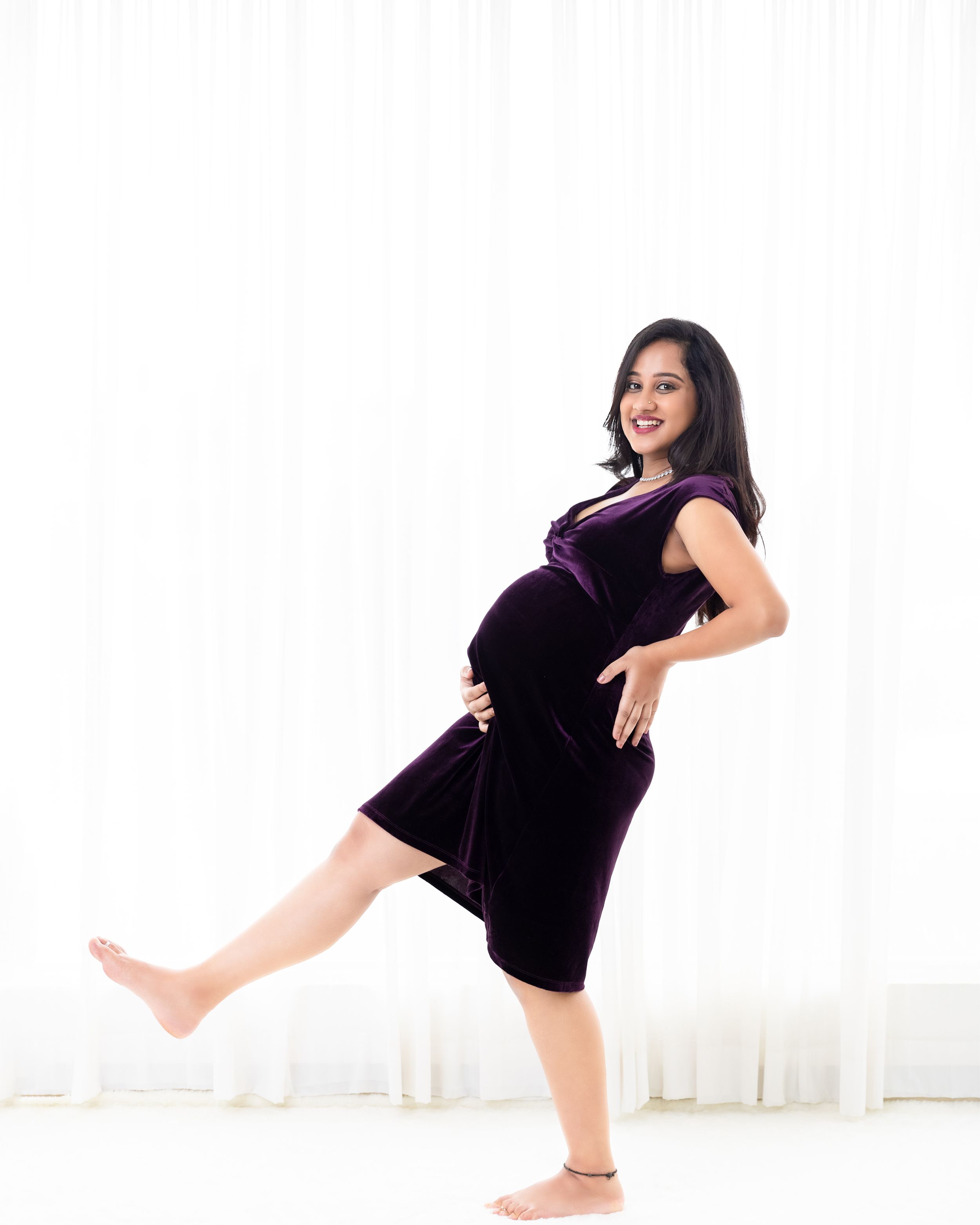 A maternity photo of someone in a purple velvet dress striking a playful pose against a white backdrop.