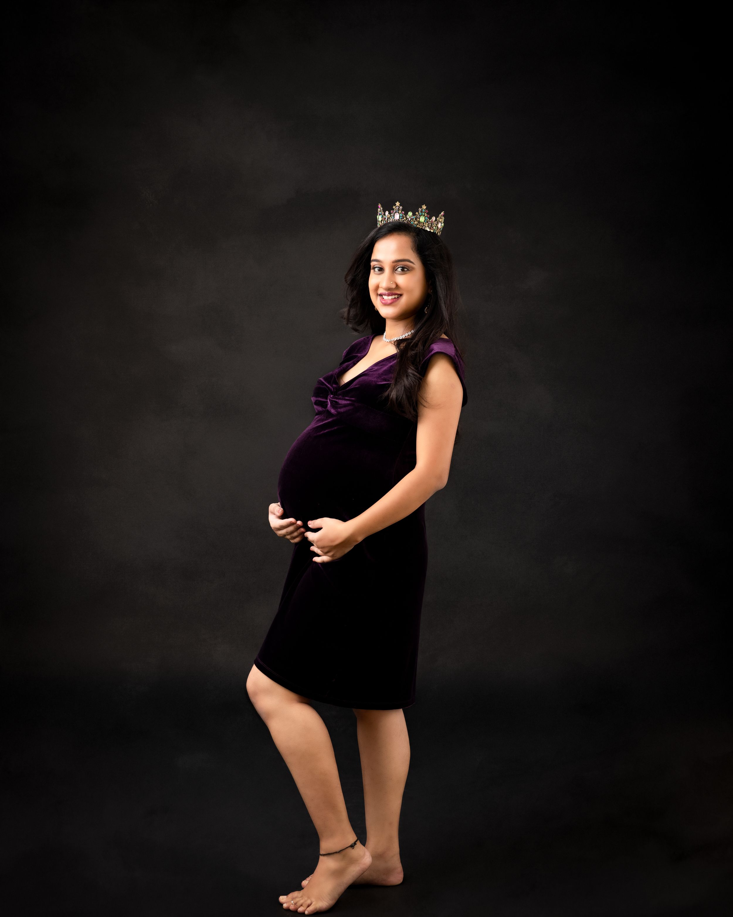 A regal maternity portrait with someone wearing a crown and purple velvet dress against dark background.