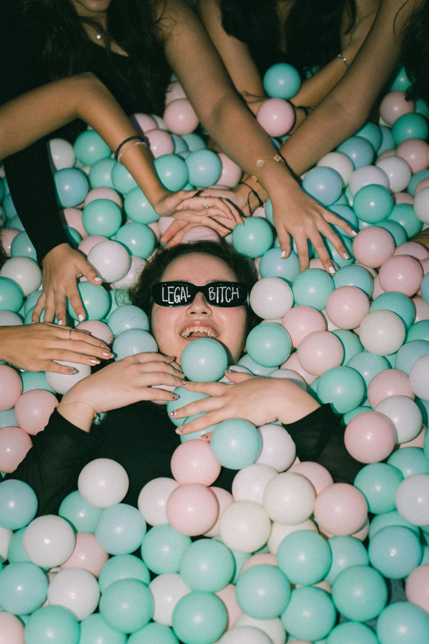 Person lying in a ball pit filled with pink and turquoise plastic balls while friends place hands on them.