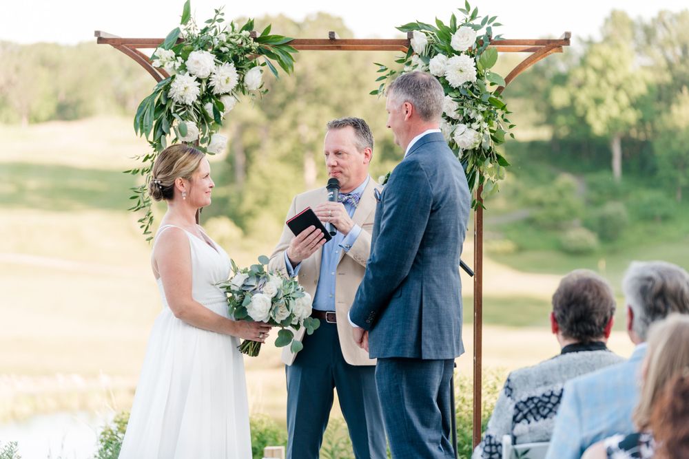 A couple exchanges vows during a sunset ceremony under a floral-adorned wooden arch.