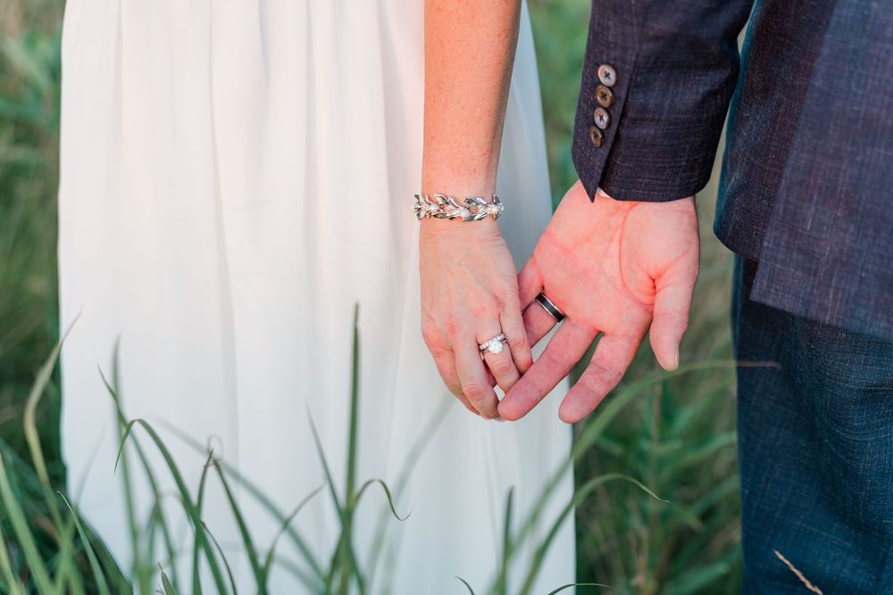 Close up of joined hands with wedding rings against white dress and natural grass background.
