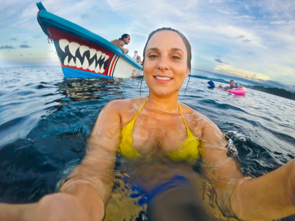 Person takes selfie while swimming with decorative boat in tropical waters.