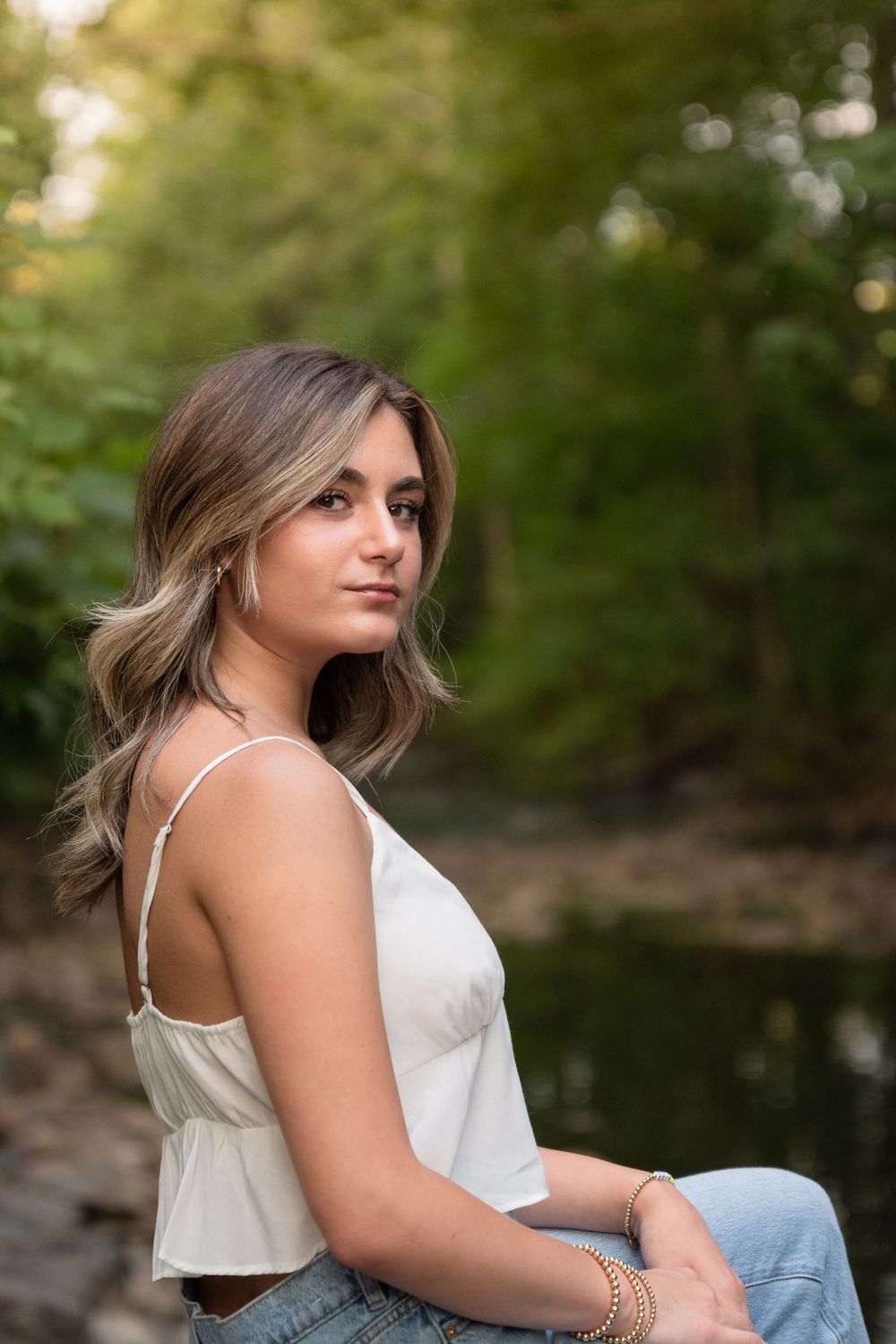 A person poses outdoors in a white top and jeans against a natural green background.