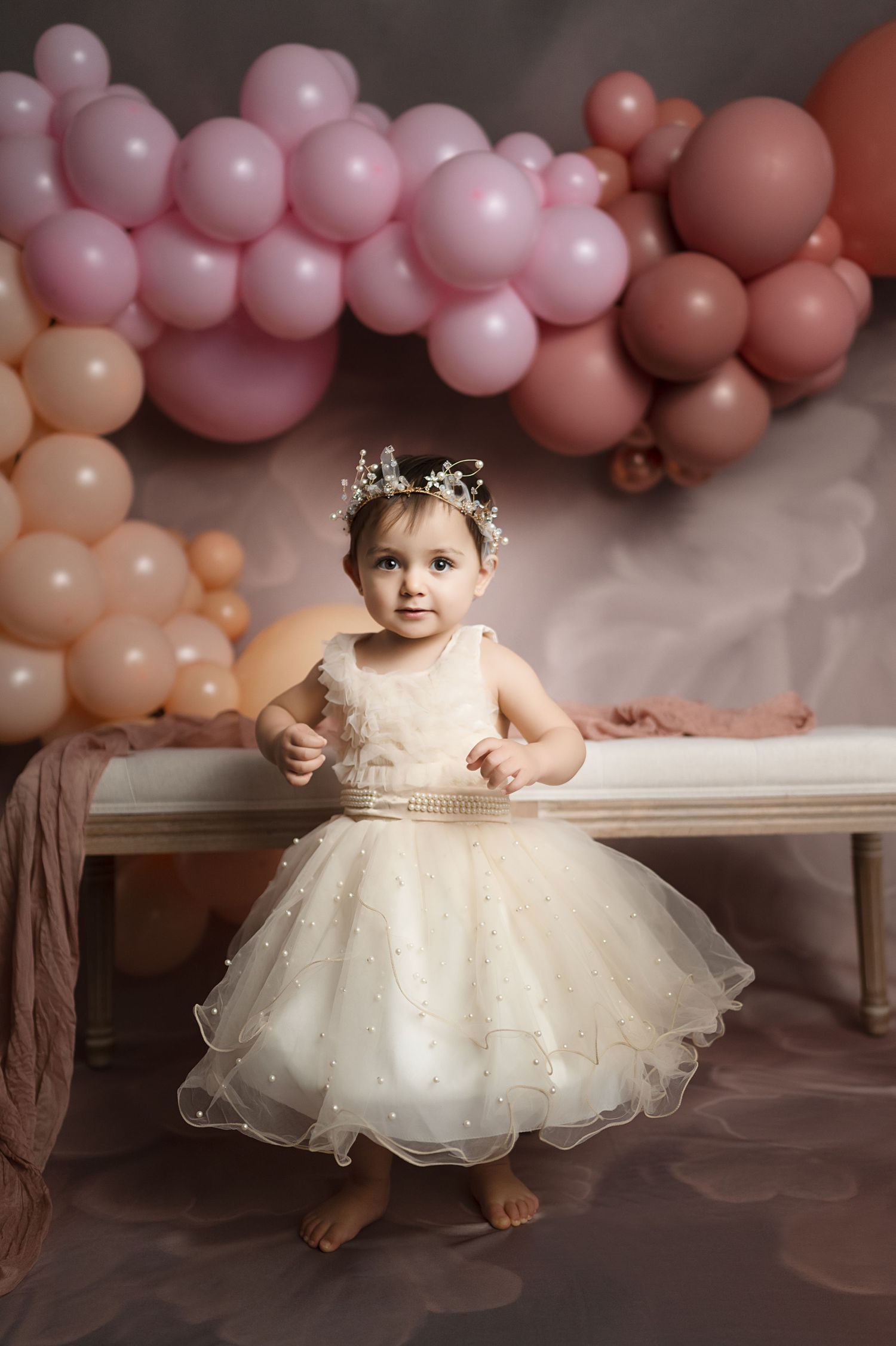 Baby in white tulle dress seated against pink balloon backdrop for first birthday photos.