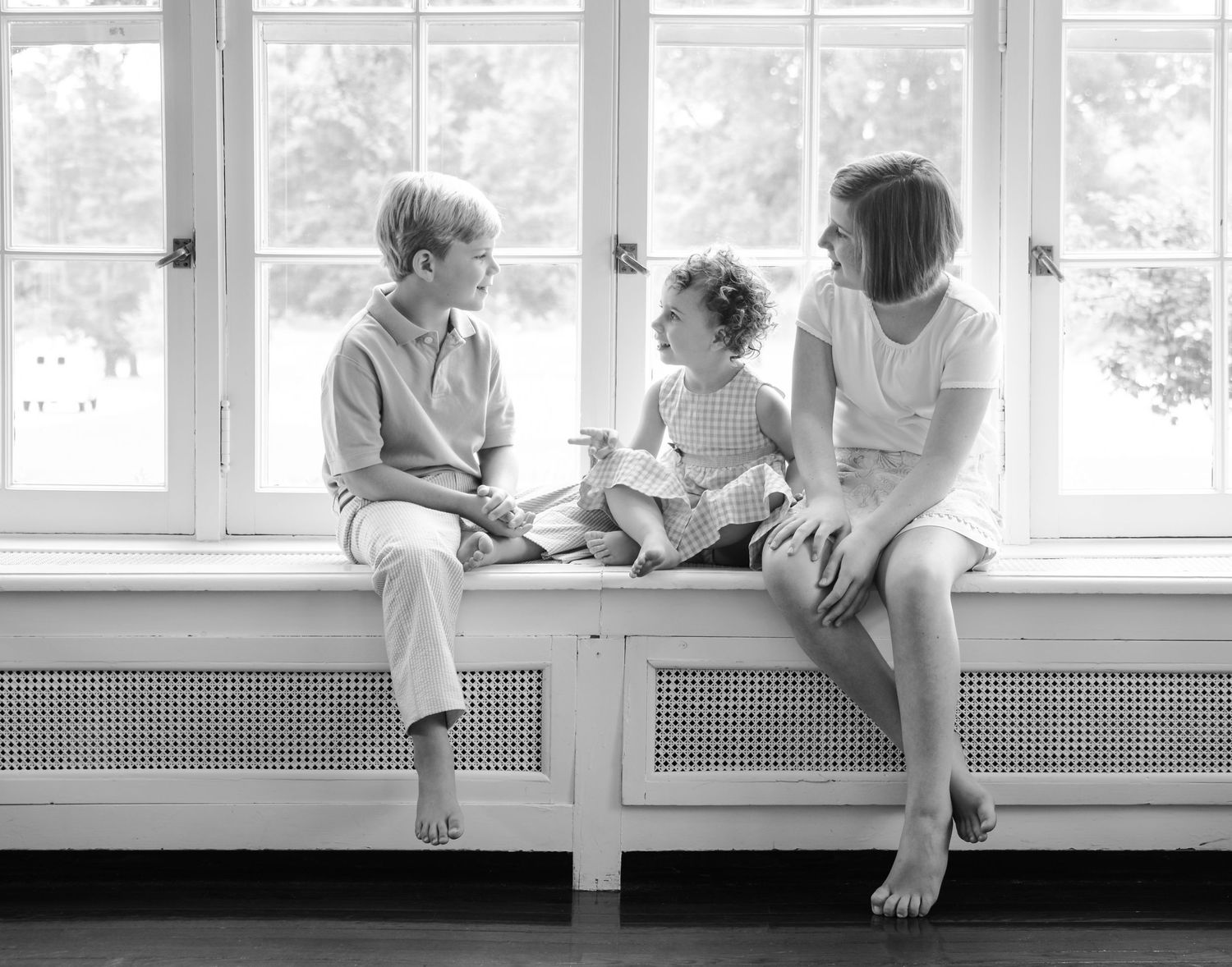 Black and white photo of three children sitting on window bench.