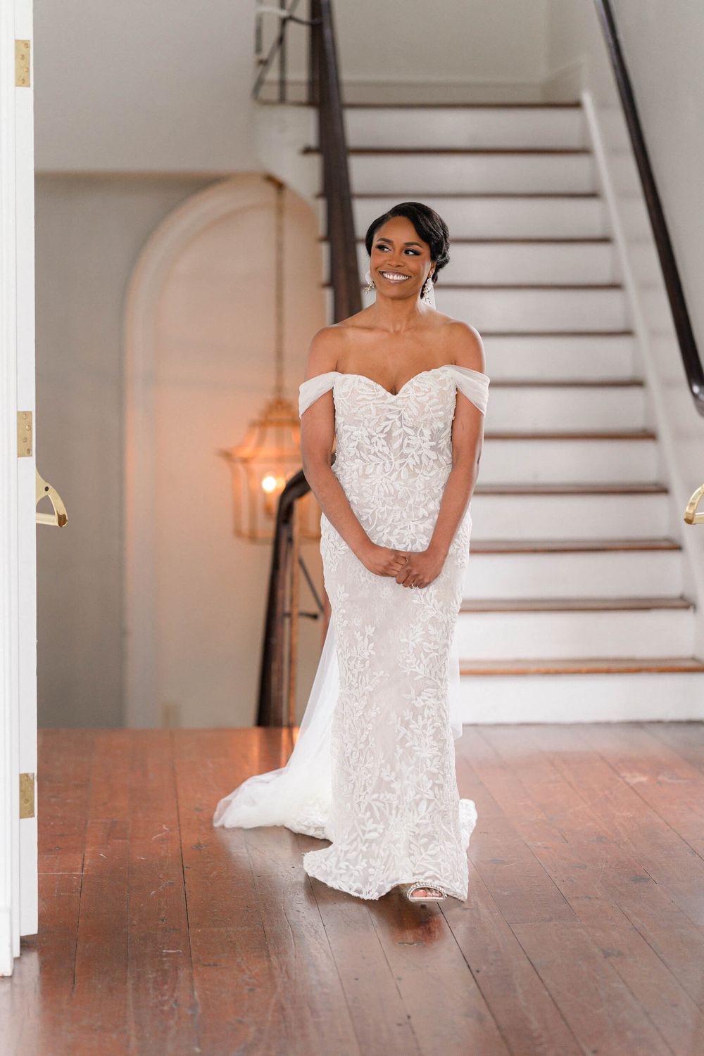 A bride poses in an off-shoulder lace wedding gown at the bottom of an elegant staircase.