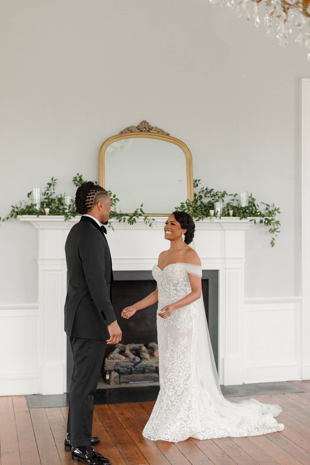 Bride and groom share a intimate moment in front of an ornate gold mirror and white fireplace with greenery.